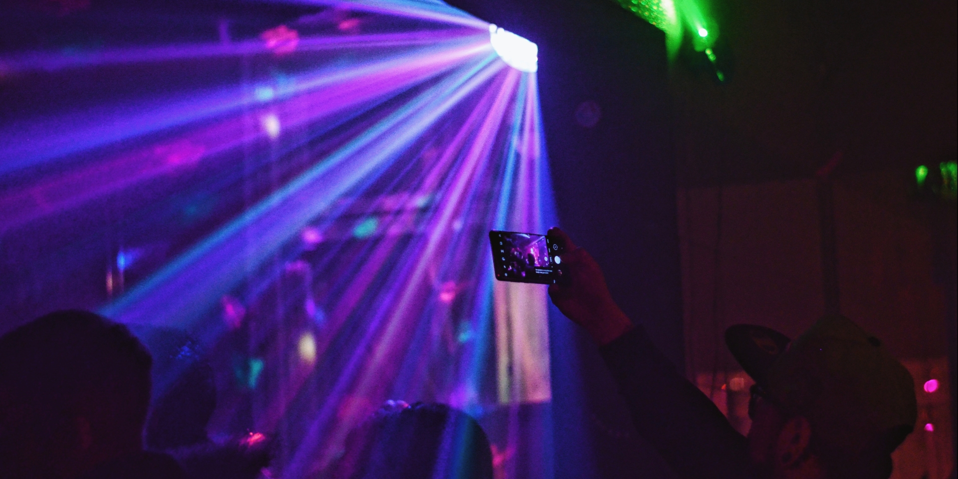 Purple strobe lights cut through a dark club. Someone stands recording on their phone. 