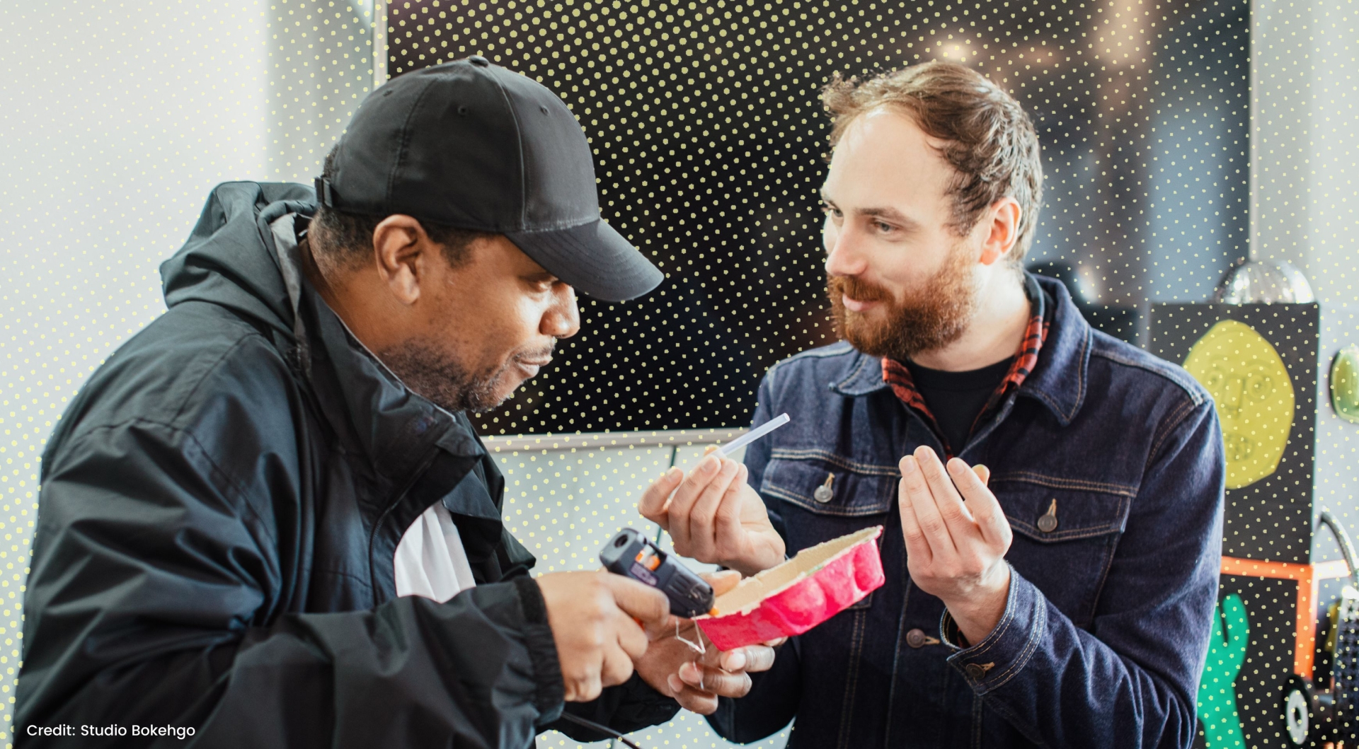 Two men sit side by side, one is using a glue gun on an egg carton whilst the other holds a glue stick