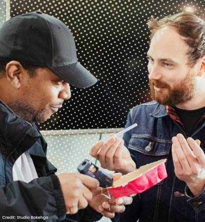 Two men sit side by side, one is using a glue gun on an egg carton whilst the other holds a glue stick